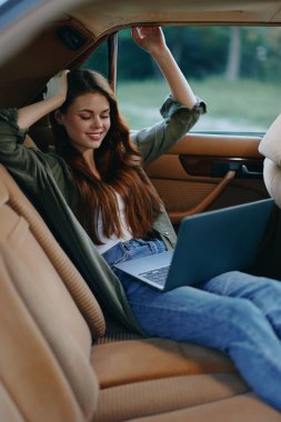 Arabadaki dizüstü bilgisayarında çalışmaktan zevk alan genç bir kadın rahat bir kıyafet giymiş, doğa ve sıcak tonlarla çevrili neşeli bir ifade sergiliyor..