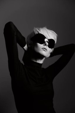 Kısa saçlı şık sarışın kadın siyah bir elbise ve büyük güneş gözlükleriyle poz veriyor. Minimalist siyah ve beyaz bir ortama güven saçıyor..