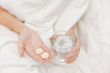 Beyaz çarşafın üzerinde yatarken elinde bir bardak su ve hap tutan kişi. resimde yatakta ilaç alan bir kişi görülüyor..