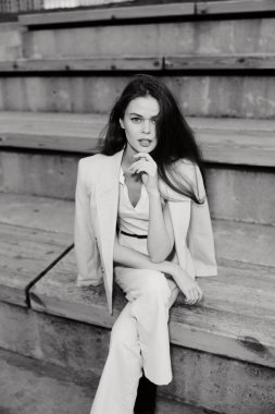 Beyaz takım elbiseli, kendine güvenen bir kadının siyah-beyaz fotoğrafı ahşap merdivenlerde oturmuş, kentli moda tarzında güçlü duyguları ifade eden siyah-beyaz iş kadını.