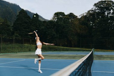 Atletik kadın, parlak mavi bir kortta tutkulu bir şekilde tenis topuna hizmet eder. Enerji ve coşku saçar. Beyaz elbisesi ağaçların yeşil arka planıyla çok uyumsuzdur.