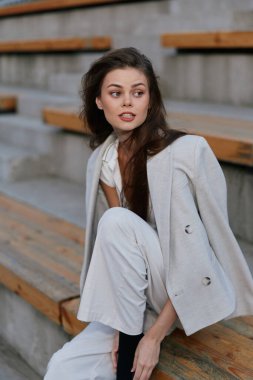 Açık bej takım elbiseli düşünceli genç bir kadın ahşap açık hava tribünlerinde oturur. Sıcak sonbahar tonları ve şehir ayarlarıyla sıradan moda tarzında ince duygular ifade eder.