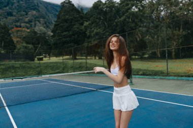 Beyaz spor kıyafetli neşeli bir bayan tenisçi, bereketli ağaçlar ve mavi gökyüzüyle çevrili, parlak, yeşil bir tenis kortunda oynarken gülümsüyor.