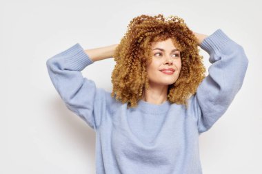 Kıvırcık saçlı, gülümseyen bir kadın açık mavi bir süveter içinde, minimalist beyaz bir arka plana karşı neşeli ifadesi ve rahat duruşu doğal güzellik ve güven duygusunu iletir.