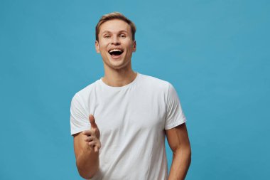 Mutlu, beyaz tişört giyen, mavi arka planda duran, ağzı açık neşeli bir yüz ifadesi ve stüdyo resminde kahkahalar gösteren mutlu bir adam. Konsept: pozitif duygu ve