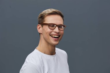 Gözlüklü, beyaz tişörtlü, akıllı telefonlu, gülen genç beyaz bir adam dijital sohbetin keyfini çıkarıyor, zihinsel sağlığı ve dijital detoksifikasyonu destekliyor. Gri arka planda izole edilmiş stüdyo portresi.