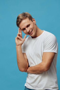 Beyaz tişört giyen gülümseyen genç adam mavi arka planda pozitif duygu, günlük tarz, zihinsel sağlık ve araç gereç farkındalığı gösteriyor. Odaklanmış modern yaşam tarzı kavramı