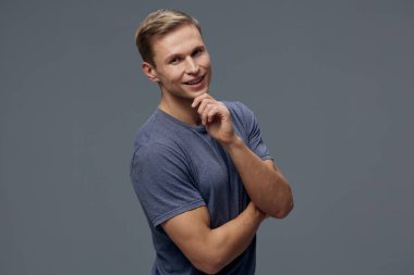 Mavi tişörtlü, gülümseyen beyaz bir adam çenesine yakın düşünerek poz veriyor. Gri arkaplanda izole edilmiş kameraya bakın. Rahat giyimli ve kısa boylu, kendinden emin dijital göçebe çalışma konsepti