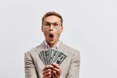 Şaşırtıcı genç beyaz adam dolar banknotlarını elinde tutuyor bej ceket ve gözlük takıyor. Gri arka planda heyecan ve para başarısı kavramını gösteriyor.