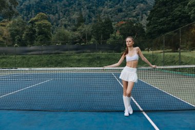 Spor kıyafeti giymiş genç bir bayan atlet, yeşil dağlarla çevrili bir tenis kortunda poz verirken kendine güveniyor. Parlak, canlı bir ortam sağlıklı bir yaşam tarzını vurguluyor.