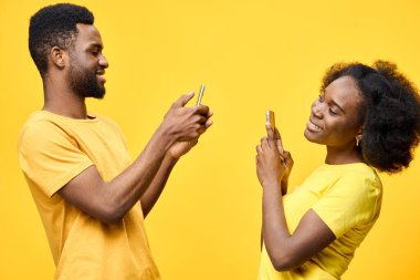Neşeli genç bir Afrikalı çift akıllı telefonlarıyla etkileşime girer, parlak sarı bir arka plana mutluluk yayar, modern gündelik giyim ve bağlantıyı vurgular