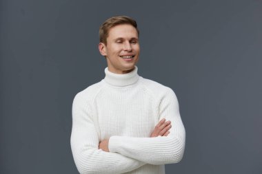 Beyaz süveter giyen gülümseyen adam kollarını çaprazladı. Neşeli, kendinden emin, beyaz tenli genç bir adam. Gri arka planda çekilen günlük kıyafet stüdyosu dijital detoks ve akıl sağlığını temsil ediyor.