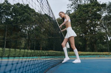 Beyaz tenis kıyafeti giymiş genç bir atletik kadın, çevrenin yeşillik, spor, spor ve sağlık kavramları ile çevrili olduğu tenis kortuna güvenerek ağın yanında duruyor.