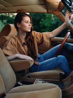 Uzun saçlı, bej gömlekli, mavi kotlu şık bir kadın klasik bir arabanın içinde dinleniyor. Macera, özgürlük ve nostalji konuları için mükemmel..