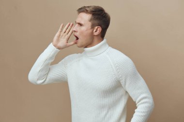 Beyaz örgü süveter giyen beyaz tenli genç bir adamın bej arka planda ağzının yanında bağırışının yan profili. Serbest bırakılma ve iletişim duyguları, zihinsel sağlık ve dijital detoks teması