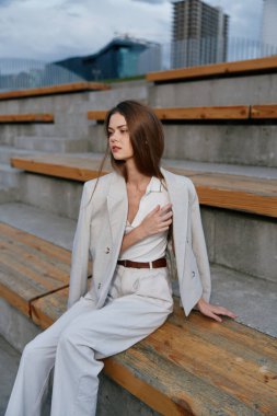 Ahşap basamaklarda oturan, modern şehir ortamında özgüven ve entelektüelliği somutlaştıran, açık renkli takım elbiseli, dengeli bir kadın, iş teması için mükemmel..