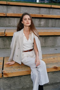 Kendine güvenen, bej renkli iş kıyafeti giyen genç Avrupalı kadın ahşap şehir merdivenlerinde oturuyor. Tarz sahibi, profesyonel görünümlü, uzun kahverengi saçlı, doğal makyajlı, modern iş modasında..