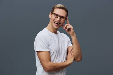 Gözlüklü, beyaz tişörtlü, gri arka planda tek başına duran gülümseyen adam parmaklarıyla dokunarak kameraya bakan neşeli dijital göçebe teknolojisini gösteriyor.
