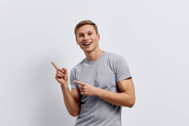 Gülümseyen, gri tişörtlü, mutlu, beyaz bir adam. Aydınlık arka planda parmaklarını heyecanlı bir şekilde işaret ediyor. Olumlu duyguları olan neşeli adam dijital detoks ve akıl sağlığını destekliyor.