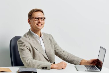 Dizüstü bilgisayarda çalışan, gözlük takan ve iş kıyafeti giyen gülümseyen bir adam, beyaz arka planda modern çalışma alanı olan bir sandalyede rahat bir şekilde oturuyor. Serbest çalışan dijital göçebe kavramı