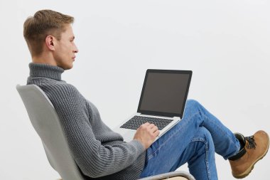 Yan görüşlü genç, beyaz arka plan teknolojisi izole edilmiş, gri süveter ve mavi kot pantolon giyen, dizüstü bilgisayarda çalışan, beyaz arka plan teknolojisi olan bir beyaz adam. Uzak çalışma, internet