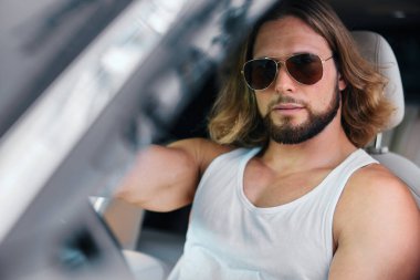Uzun saçlı, güneş gözlüklü şık bir adam bir arabanın şoför koltuğunda rahatlar. Kendine güveni ve zarafeti vardır.