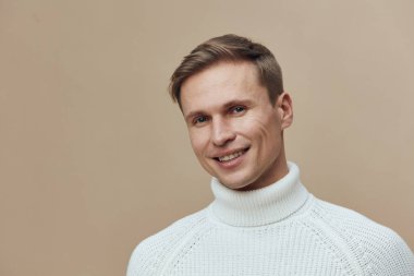 Beyaz balıkçı yaka kazak giyen, kısa saçlı gülümseyen adam bej arka planda izole edilmiş kameraya bakıyor. Beyaz adamın portresi dijital göçebe ve uzak çalışma yaşam tarzını temsil ediyor.