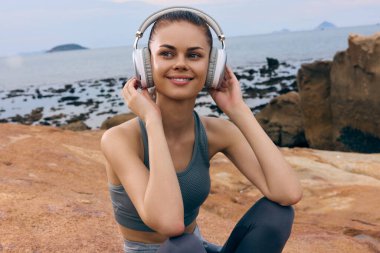 Kulaklık takan mutlu genç bir kadın, spor kıyafeti giymiş, sakin sahil atmosferinin tadını çıkarıyor. Müzik, rahatlama ve açık hava aktiviteleri için mükemmel.