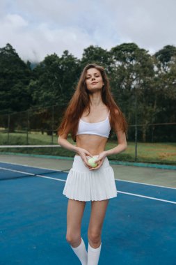 Beyaz spor kıyafetli enerjik genç bir kadın, mavi kortta bir tenis topu tutuyor, yeşil ağaçlarla çevrili, fitness ve atletizmi sembolize ediyor.