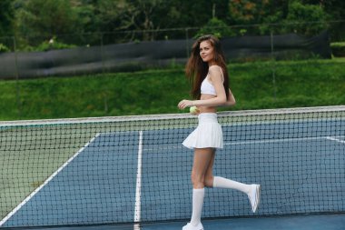 Tenis kortunda beyaz bir spor kıyafetiyle gülümseyen genç bir kadın, enerji ve neşe saçıyor, aktif bir yaşam tarzını ve atletizmi temsil ediyor.