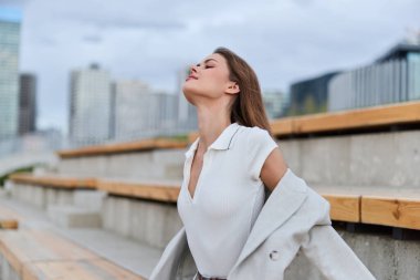Beyaz bluzlu ve gri ceketli genç bir kadın şehir ortamında sakin bir şekilde kendine güveni kapalı gözlerle, gündelik moda ve sonbahar havasını yansıtıyor.