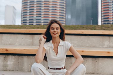 İş hayatında kendine güvenen bir kadın, açık havada, canlı bir şehir ortamında otururken gülümser ve kendine güvenini ve profesyonelliğini sergiler. Şehir sahnesi onun motivasyonlu ifadesini arttırıyor ve...