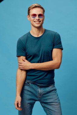 Güneş gözlüğü takan gülümseyen genç bir adam ve kollarını mavi arka plana yaslamış, pozitif duygu ve modern tarzı yansıtan, çapraz kollarıyla rahat duran bir tişört. Konsept: Ruh sağlığı ve cihazlar.