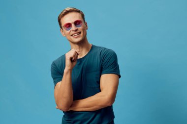 Kısa saçlı, pembe güneş gözlüklü, gülümseyen ve kameraya bakan sıradan mavi tişörtlü, sağlam mavi arka planda duran, rahat ruh hali ve pozitif titreşimli mutlu genç adam. Kavram: Neşeli