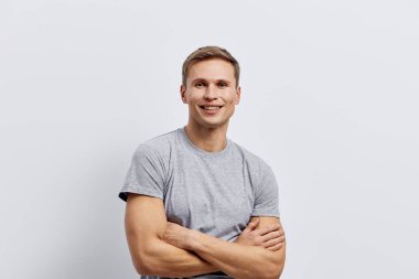 Sıradan gri tişörtleriyle kollarını kavuşturup beyaz arka planda duran kendine güvenen bir adam. Stüdyo görüntüsü neşeli, doğal ışıklandırmayla ulaşılabilir bir ruh hali yakalıyor, erkek model.