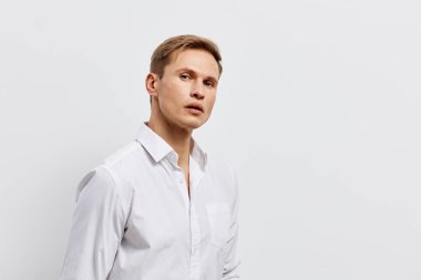 Beyaz gömlekli, açık kahverengi saçlı seçkin bir adam stüdyo ortamında beyaz bir arka plana karşı ciddi bir ifadeyle poz veriyor. Konsept: profesyonellik ve güven