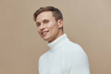 Beyaz balıkçı yaka kazaklı mutlu bir gencin portresi gülümseyen ve bej arka planda kameraya bakan. Neşeli ve arkadaş canlısı bir ruh hali, yaşam tarzı, moda veya sağlık için uygun.