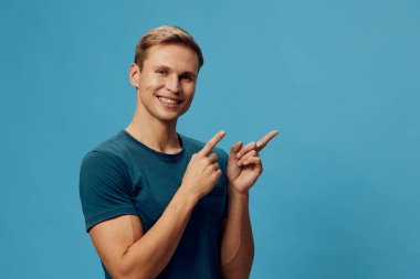 Kısa saçlı, günlük mavi tişörtlü mutlu bir genç adam, iki eliyle açık mavi bir arka plana işaret ederken geniş bir gülümseme sergiliyor. Kavram: gençlik sevinci ve pozitiflik, rahatlama