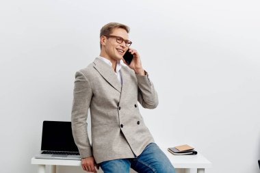 Kendine güveni tam, şık bir spor ceket giymiş bir adam telefon konuşmasına dalmış, minimalist bir ofis ortamında dizüstü bilgisayar ve not defterleriyle duruyor, parlak ve profesyonel.