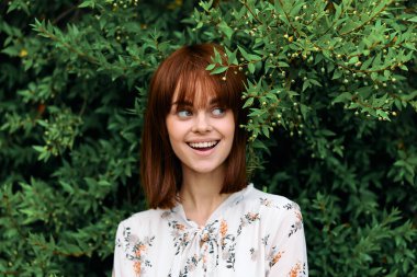 Canlı, kızıl saçlı bir kadın yemyeşil bitkilerin ve çalıların önünde poz veriyor.