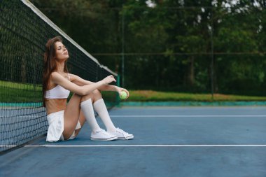 Genç atletik bir kadın, spor bir tenis kıyafeti içinde, etek ve üst ile, bir tenis kortunda gevşiyor, yeşillik, vücut bulmuş denge ve sporda rahatlıkla