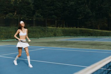 Şık beyaz tenis kıyafetli enerjik genç bir kadın canlı bir mavi kortta oyun oynuyor, yemyeşil bir ortamda mutluluk ve atletizm yayıyor.