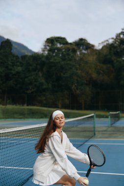 Beyaz bir tenis elbisesi içinde genç bir bayan atlet, mavi bir tenis kortunda neşe içinde oynuyor bu canlı sahne onun heyecanını ve güzel açık hava ortamını yakalıyor.