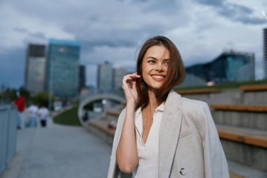 Açık bej ceket giyen kendine güvenen bir kadın şehir iş ortamında gülümsüyor. Sonbahar sezonu, şık gündelik moda, binaları olan modern şehir arka planı..