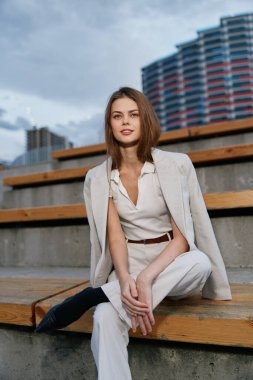 Beyaz iş elbisesi ve bej ceket giyen kendine güvenen bir kadın modern mimari ve bulutlu gökyüzü ile kentsel çevrede ahşap merdivenlerde oturarak profesyonelliği ve sakinliği ifade ediyor.