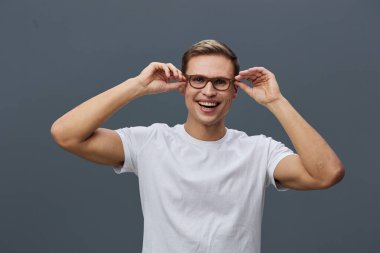 Gözlüklü ve beyaz tişörtlü mutlu beyaz bir adam koyu gri arka planda zevkle kameraya bakıyor. Günlük giysiler ve pozitif ifade güven ve rahatlığı ifade eder.