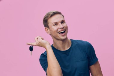 Kısa açık kahverengi saçlı neşeli bir adam, günlük mavi tişörtler giymiş, geniş bir gülümsemeyle arabanın anahtarlarını tutuyor, düz pembe bir arka planda duruyor. Kavram: Yeni başlangıçların mutluluğu