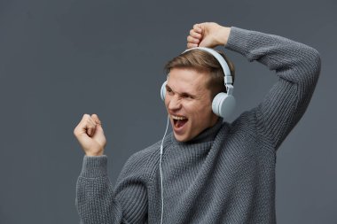 Gri kazaklı mutlu adam kulaklıklı müzikten hoşlanıyor, heyecan ve enerji gösteriyor. İzole edilmiş gri arka plan, onun neşeli ruh halini, rahat stilini, yaşam tarzını, duygularının kutlanmasını vurgular..