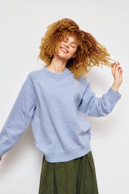 Açık mavi kazaklı, kıvırcık saçlı neşeli kadın manken anlık bir anın tadını çıkarıyor, pozitiflik ve kendine güven yayıyor. Minimalist beyaz arka plan onun neşeli havasını ve modasını artırıyor.