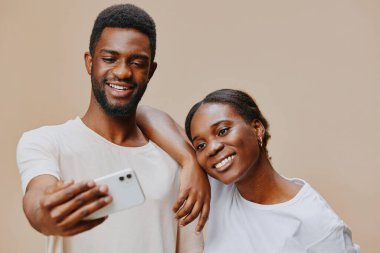 Neşeli genç bir çift, akıllı telefonlarıyla selfie çekerken, tarafsız bir arkaplan üzerine kurulmuş bu resim aşkı, mutluluğu ve modern teknolojiyi gösteriyor.